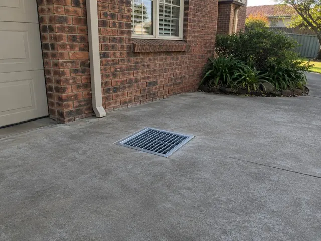 Clean outdoor drain grate set in a concrete driveway beside a brick house in Morpeth