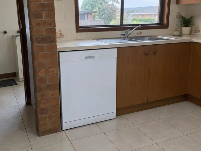 White dishwasher under a kitchen benchtop beside a sink and timber cabinets in a tiled home kitchen in Raworth