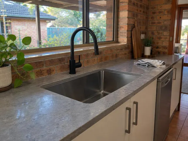 Black kitchen mixer tap over a stainless sink with stone benchtop and brick wall in Tenambit