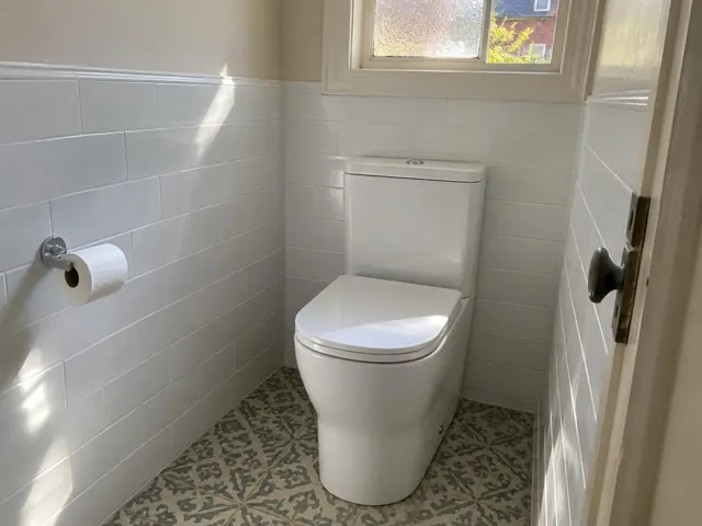 White toilet in a small tiled bathroom with patterned floor and window light in Morpeth